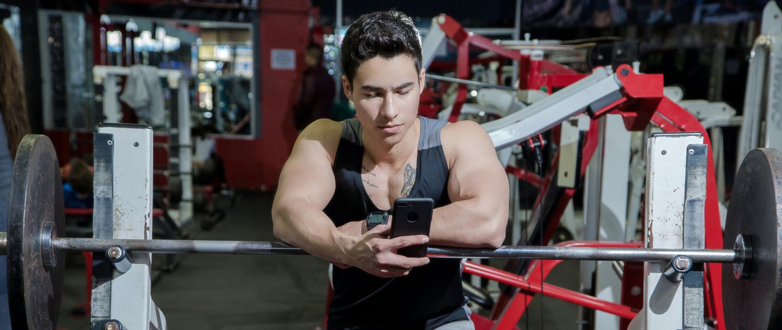 A muscular man in a tank top leans on a bar with weights at the gym, looking at his phone.