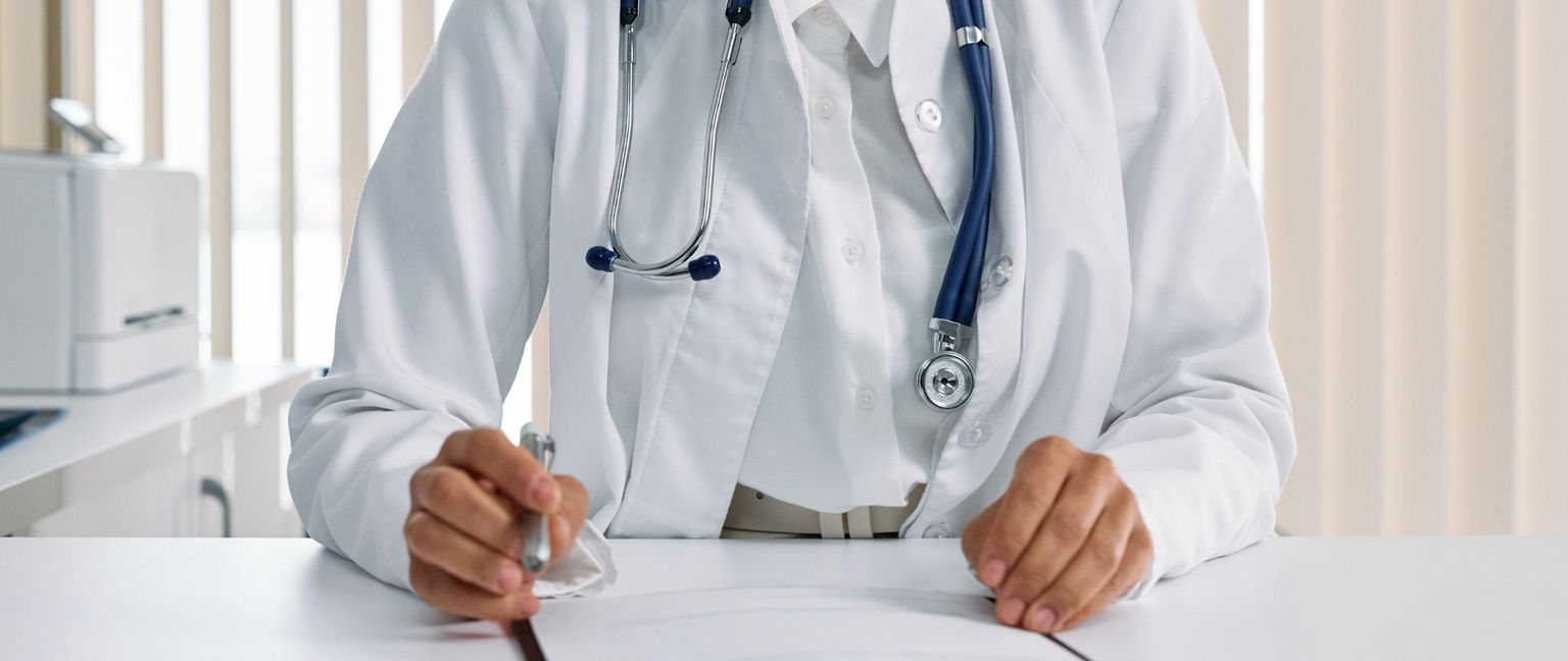 A close-up of a person in a white doctor's coat sitting at a desk with a dark blue stethoscope around their neck. They are holding a pen and writing on a document.