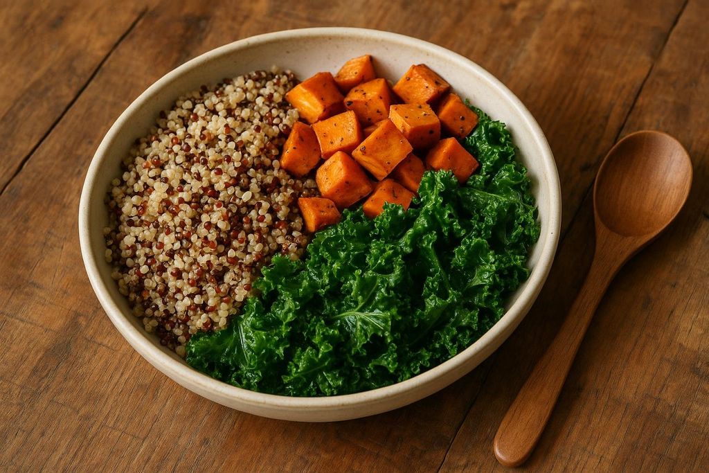A white bowl filled with three sections: fluffy quinoa, diced roasted sweet potatoes, and green kale, sitting on a wooden table next to a wooden spoon.