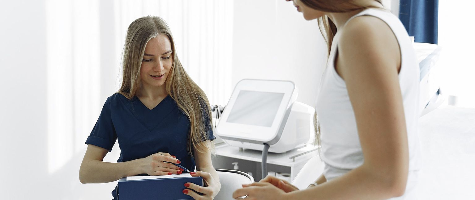 A doctor in a blue scrub top consulting with a patient wearing a white tank top, looking at a tablet together.