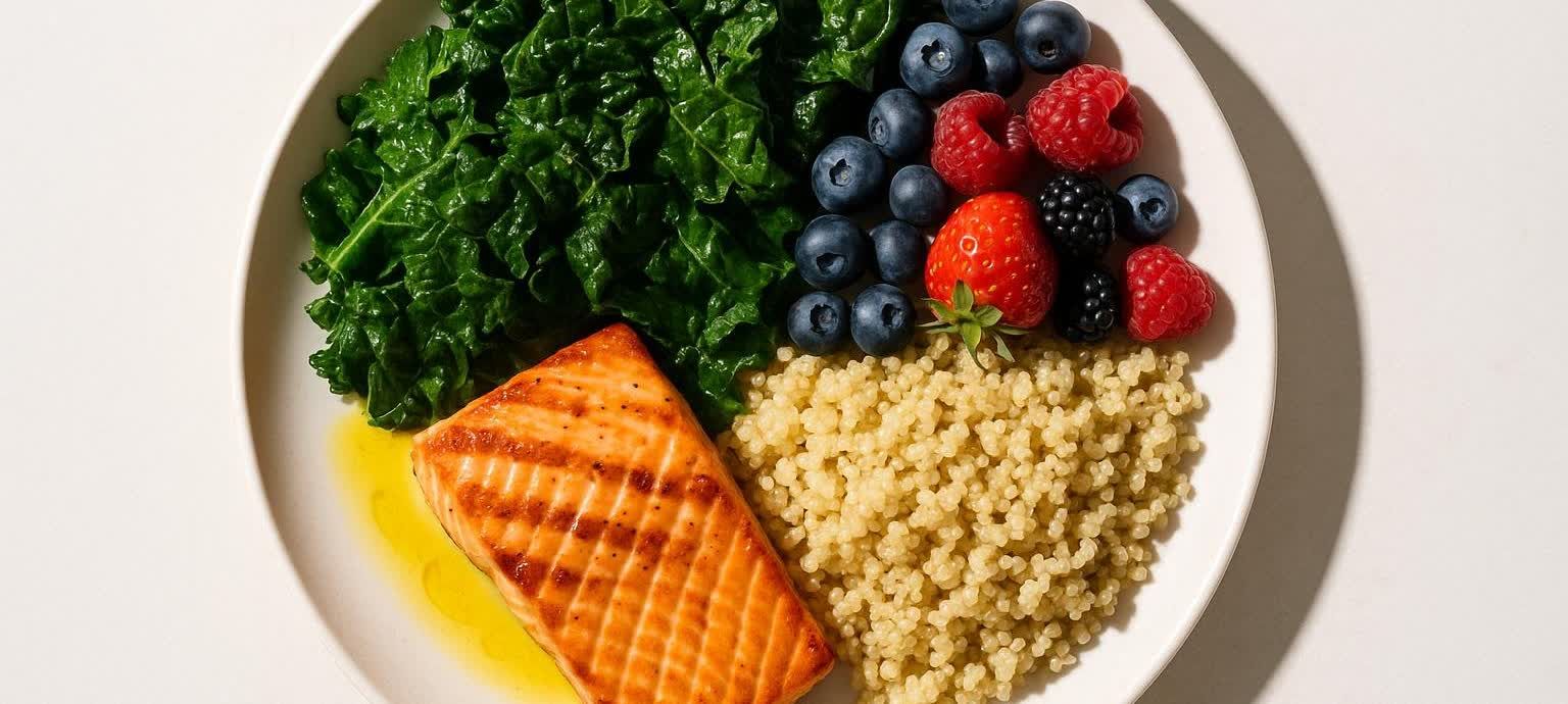 An overhead shot of a healthy and anti-inflammatory meal on a white plate. The meal includes a piece of cooked salmon, vibrant green leafy vegetables (likely kale), a portion of quinoa, and a colorful mix of berries (blueberries, raspberries, strawberries, and blackberries).