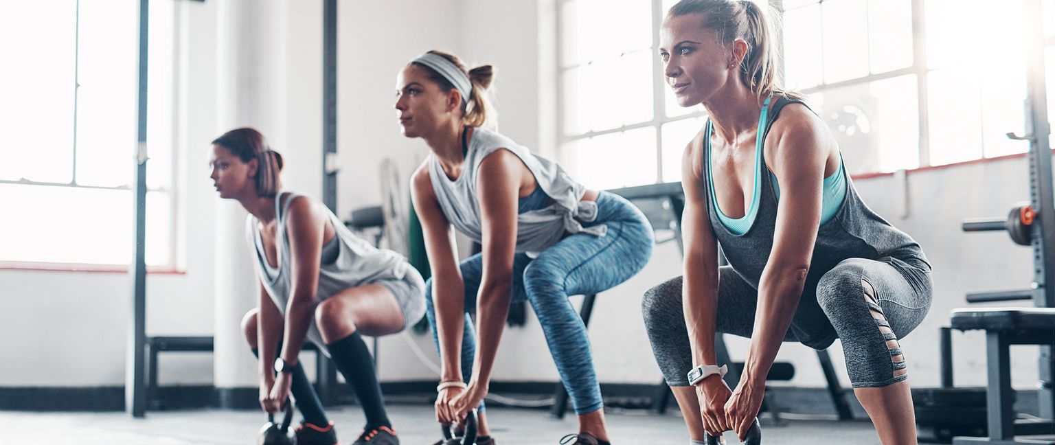 Three women performing kettlebell squats in a brightly lit gym.