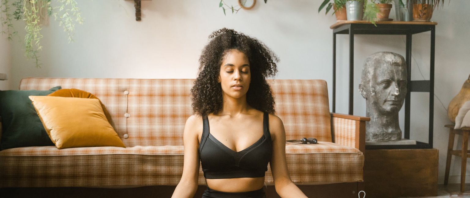 A Black woman sits in her living room with her eyes closed, meditating. Behind her is a couch with pillows and a plant, and next to her is a sculpture of a head on a stand.