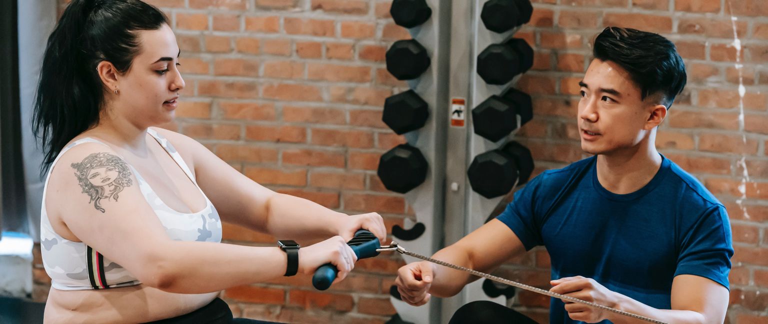 A woman with a tattoo on her arm and a man are both using a rowing machine in a gym with a brick wall and weight rack in the background.