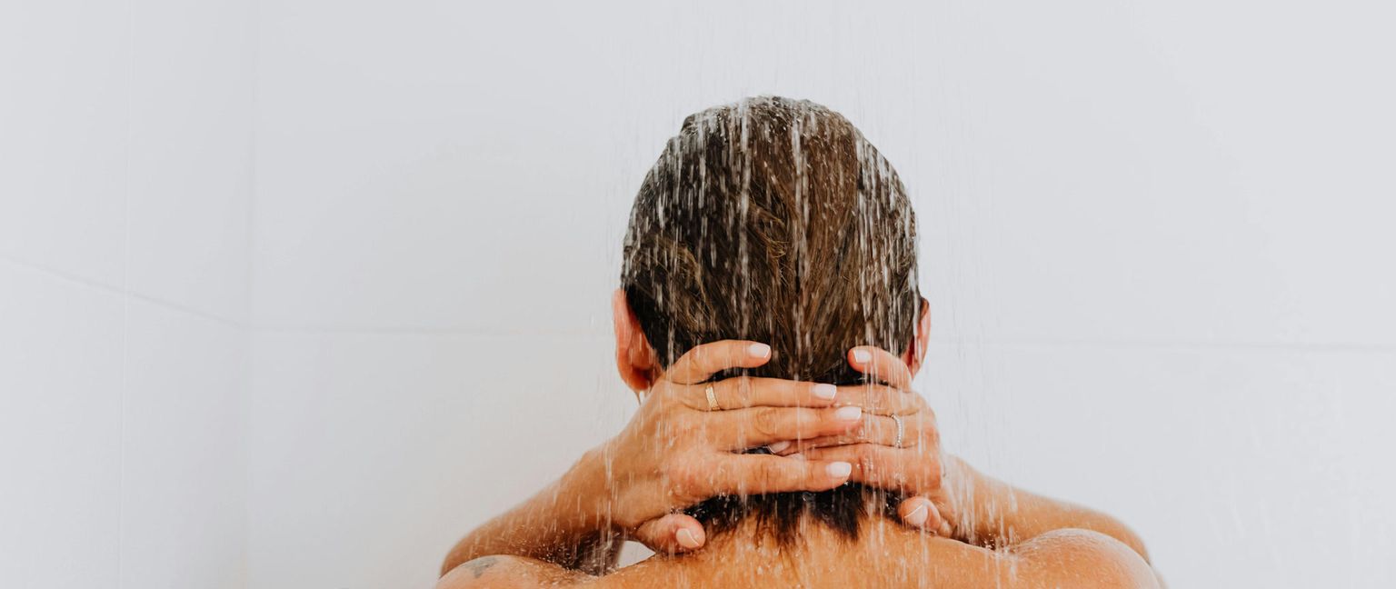 Rear view of a woman with brown hair showering. Her hands are clasped behind her neck.