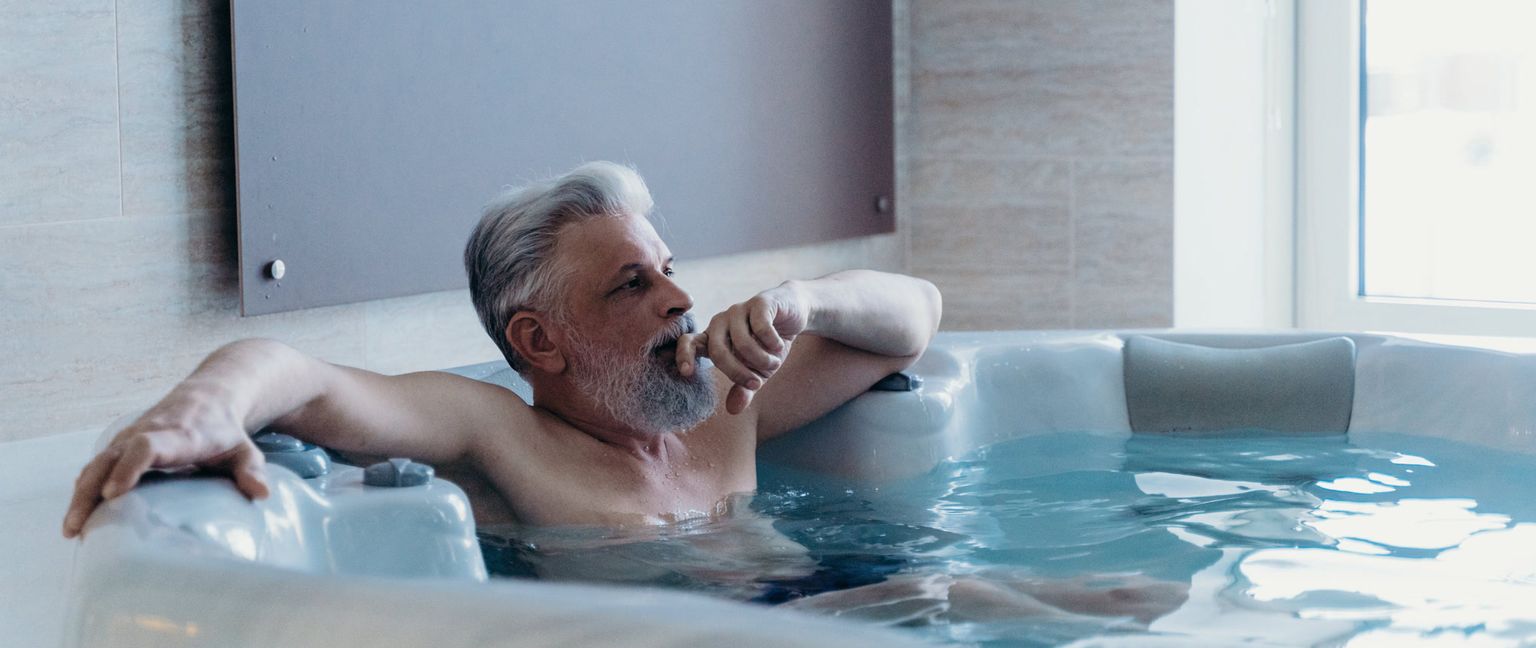 A man relaxes in a hot tub. He has gray hair and a gray beard and is looking to the side with his hand near his mouth.