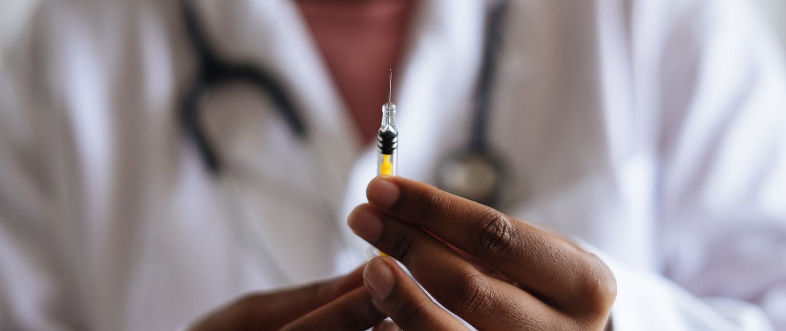 Close-up of a hand holding a medical needle against a blurry background of a white coat and stethoscope.