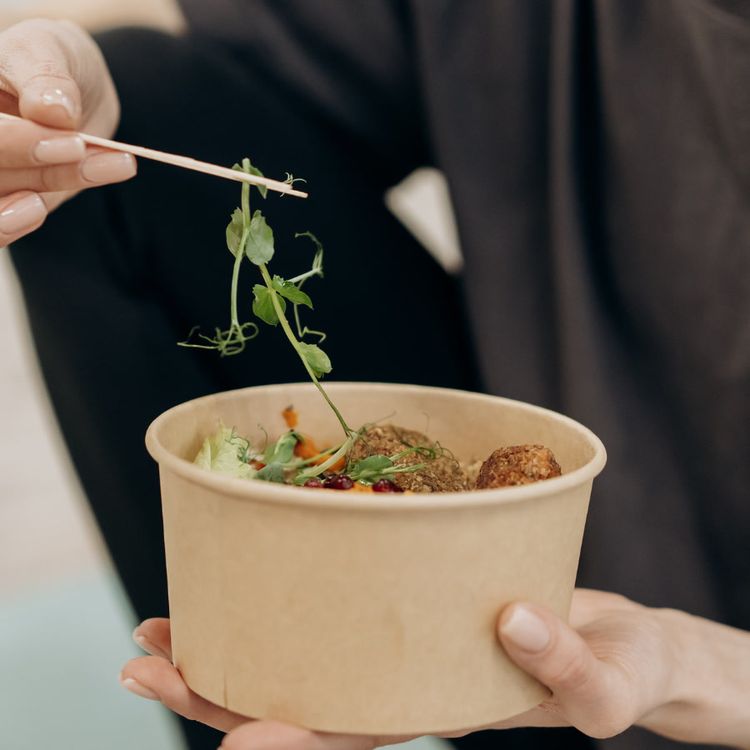 Close-up image of a person holding a disposable biodegradable food bowl filled with healthy food. Another hand is adding a sprout to the bowl with chopsticks.