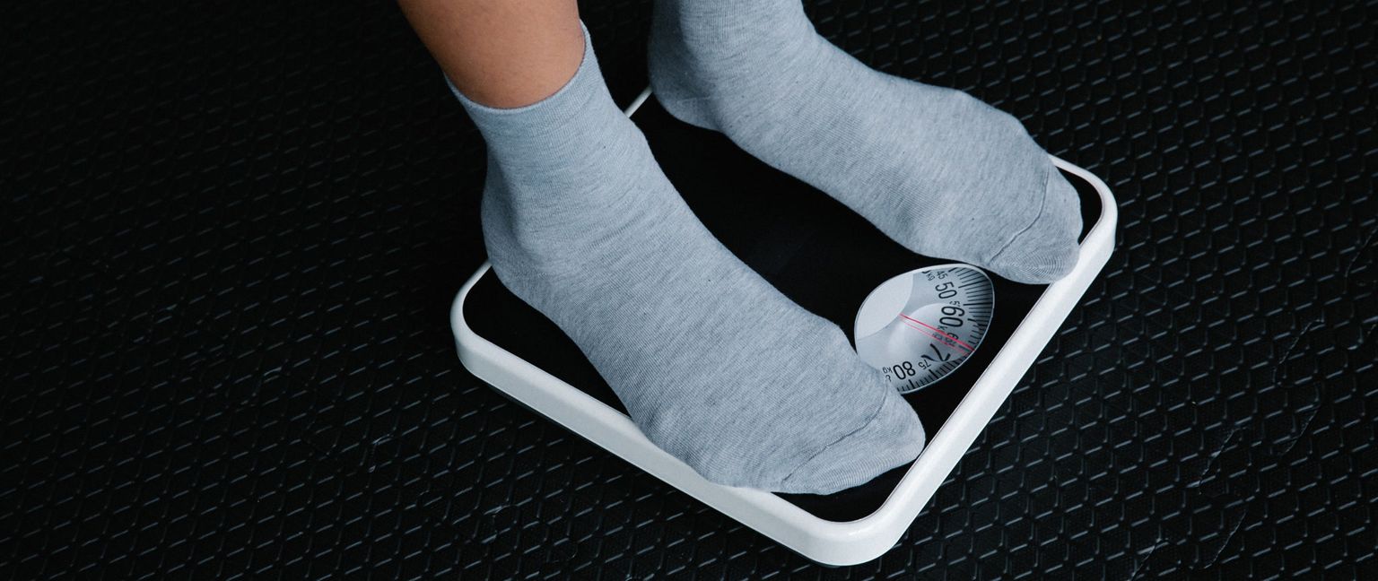 A person wearing gray socks stands on a white and black scale, with the needle pointing between 50 and 60 kilograms.