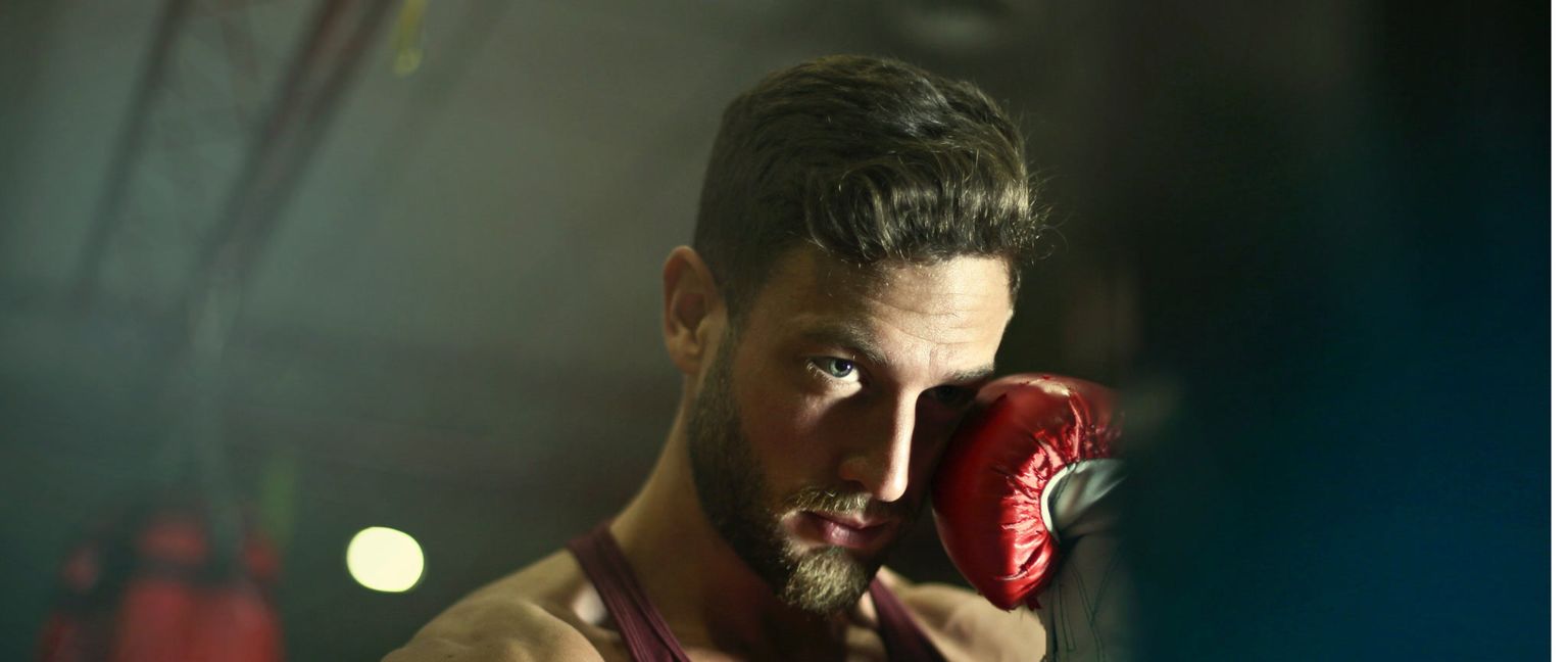 A man with a beard wearing a maroon tank top and a red boxing glove near his face looks intently forward.