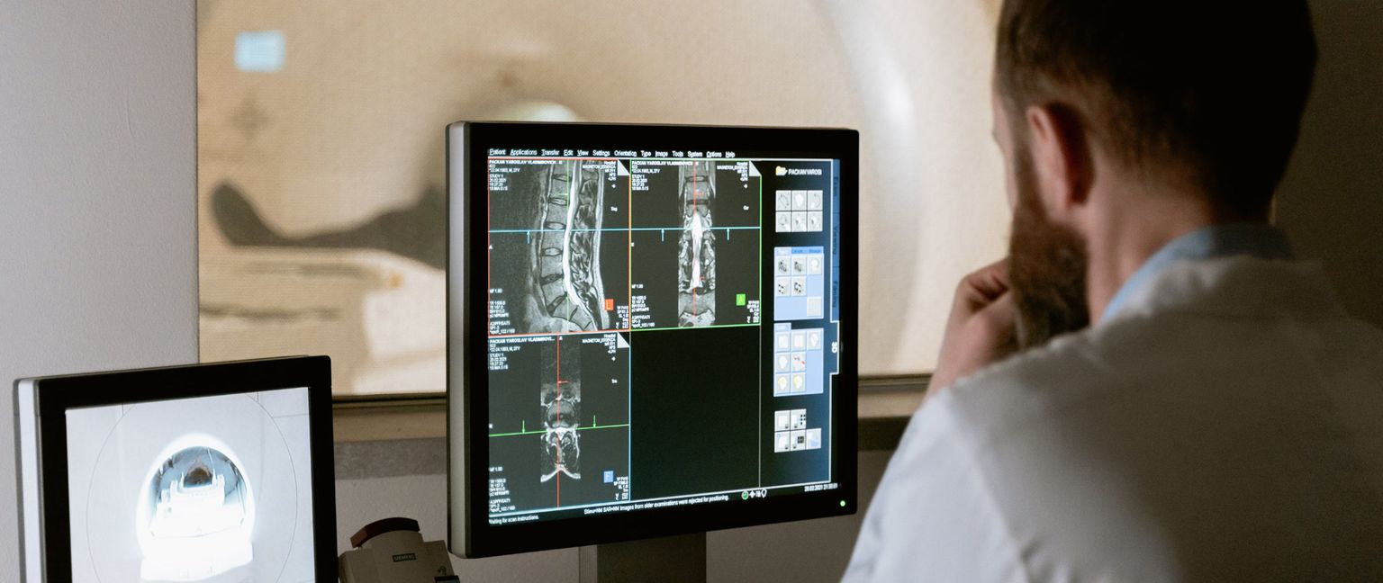 A doctor in a white coat reviews a CT scan on a computer monitor. The screen shows medical images of a spine, with text and diagrams visible from a patient's record.