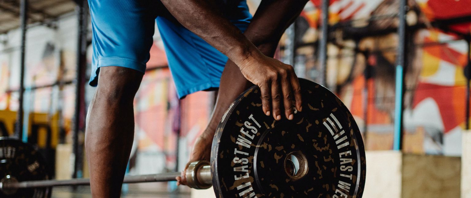 A man in blue shorts leans down to add a large, black weight plate marked "EAST WEST" to a barbell.
