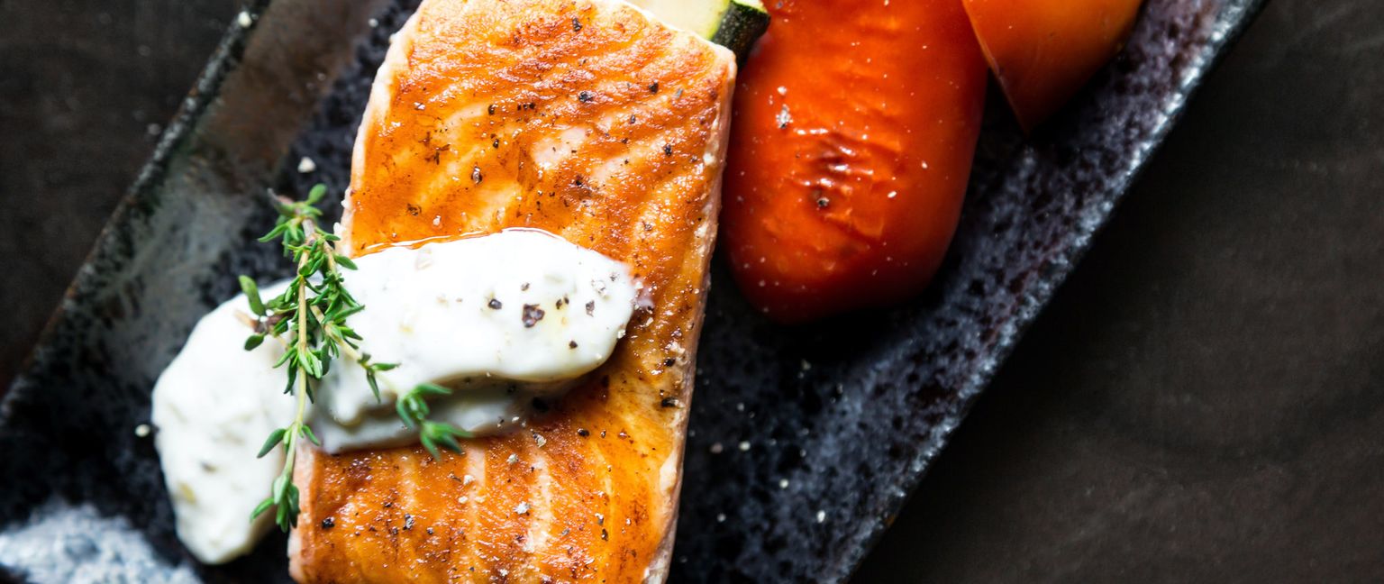 A close up shot of a pan seated salmon on a plate with cream sauce, green sprig of thyme, cherry tomatoes and slices of squash.