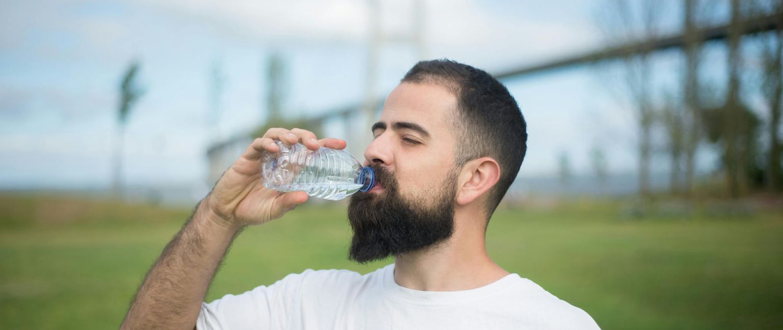 A man with a beard is drinking from a clear plastic water bottle in a park. The background is a blurred green field and blue sky with a bridge in the distance.