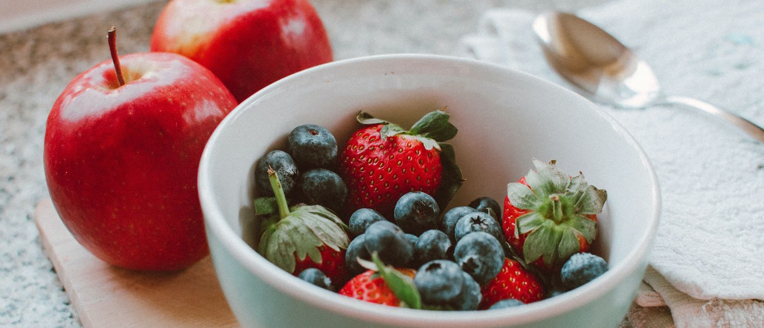 A close-up shot of a bowl of fresh strawberries and blueberries, with two red apples and a spoon in the background.