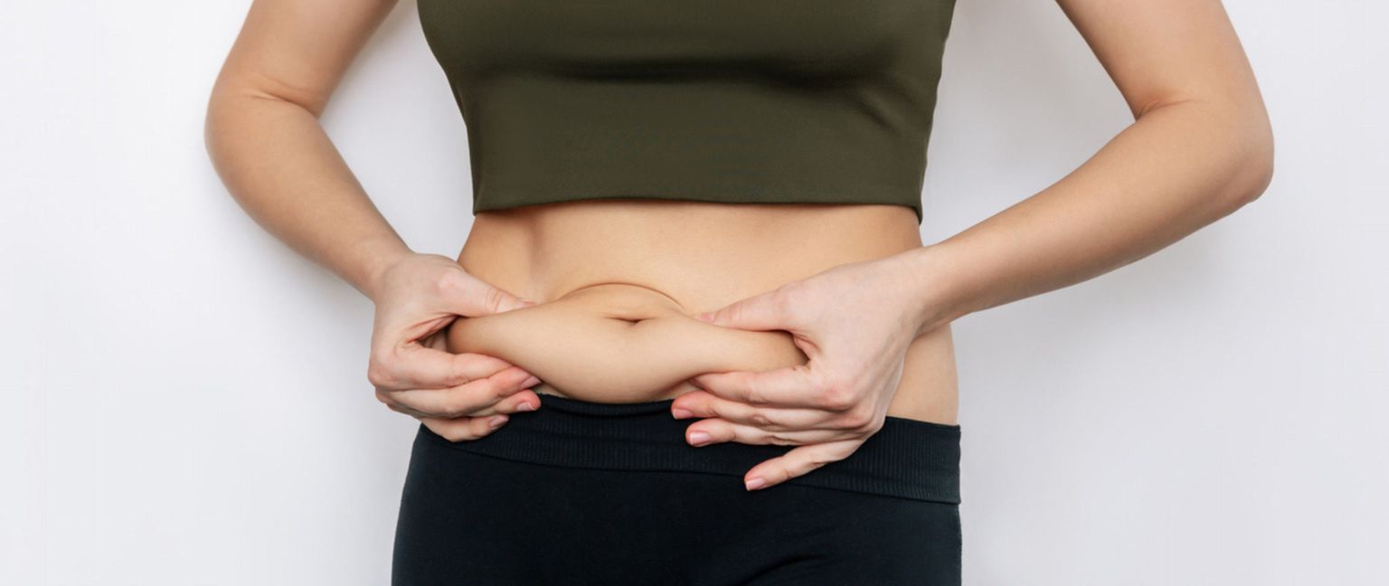 A woman in a crop top and black leggings pinches the excess skin and fat around her stomach.