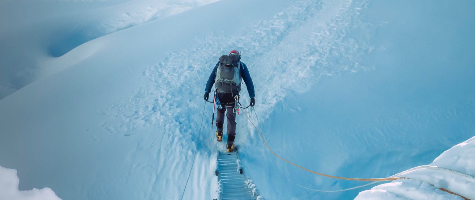 A climber on a snowy mountain path, secured with ropes, traversing a steep and icy slope.