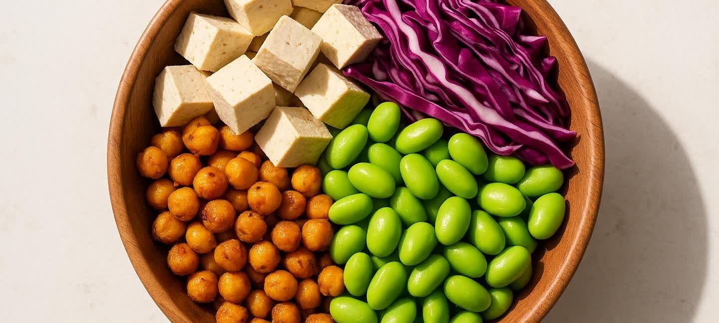 A wooden bowl filled with various vegan protein foods, including cubed tofu, roasted chickpeas, bright green edamame, and shredded red cabbage.