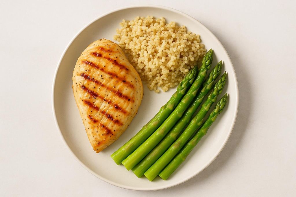A top-down view of a white plate holding a serving of grilled chicken breast with grill marks, a pile of cooked quinoa, and several vibrant green asparagus spears arranged neatly. The plate is set against a plain, light background.