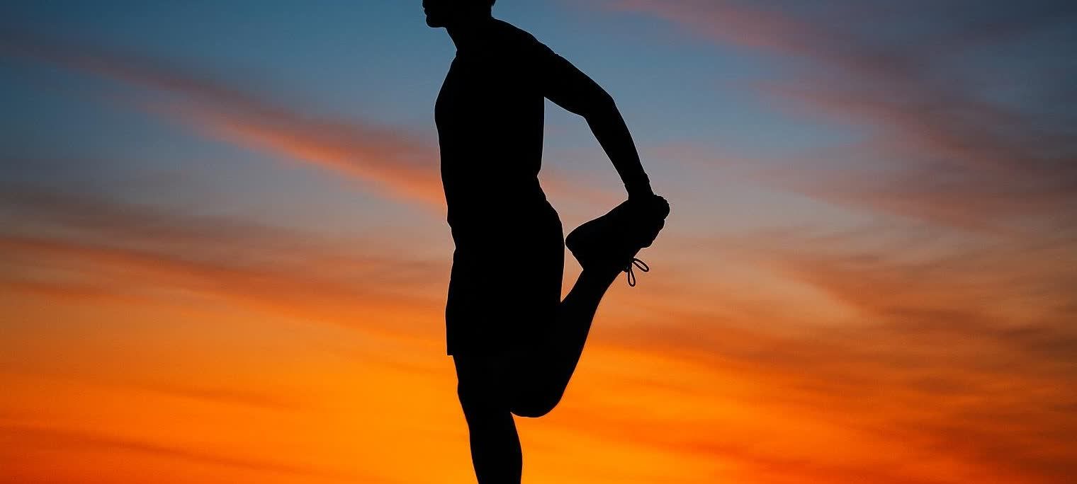 A silhouette of a runner stretching their quadriceps against a vibrant sunrise backdrop. The sky transitions from dark blue at the top to bright orange and red at the bottom, with faint clouds.