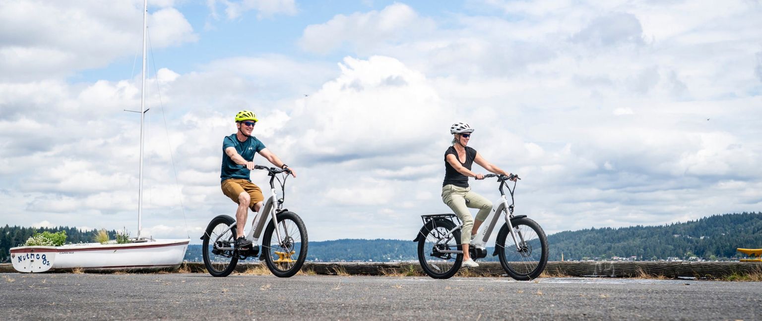 A man and woman ride electric bikes on a paved path along the waterfront, with a sailboat and blue water in the background.