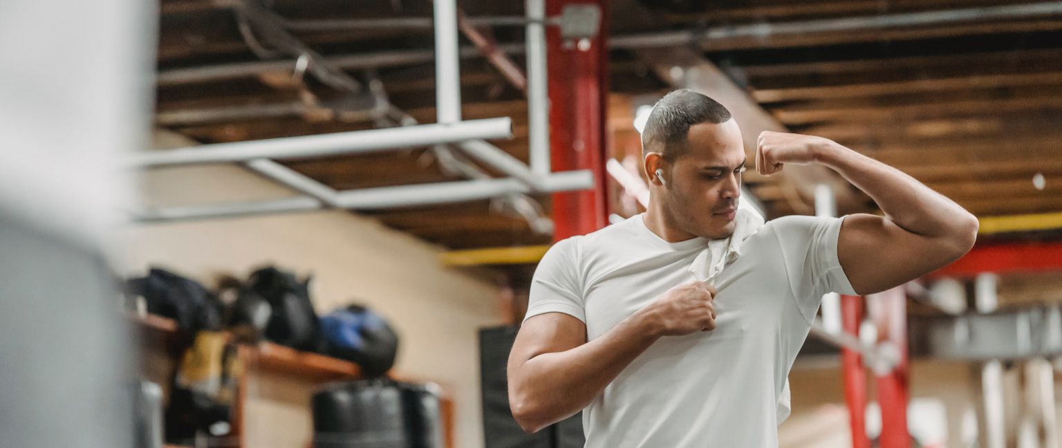 A man in a gym pauses his workout to flex his bicep. He is wearing a white shirt and has wireless headphones in his ears.