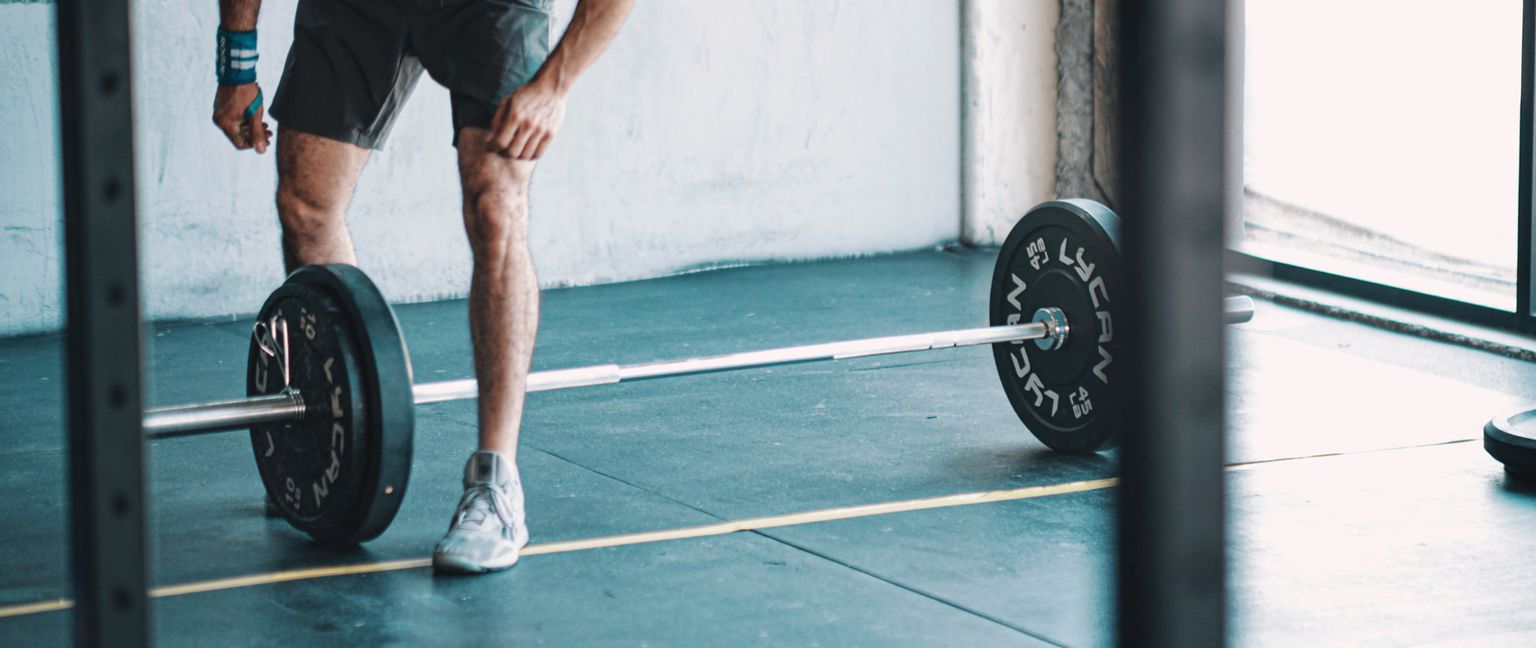 A man in fitness attire stands over a barbell with weights in a gym setting, preparing for a lift.