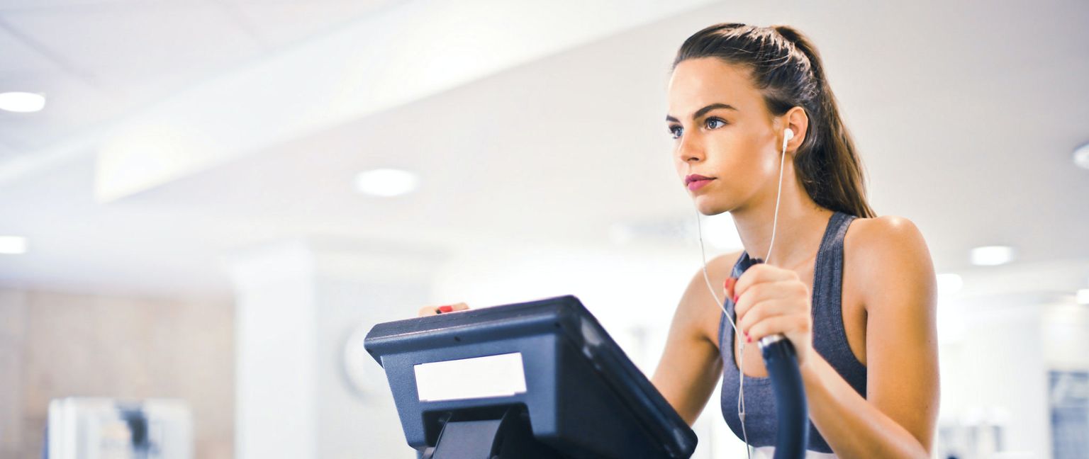 A focused woman works out on an elliptical machine in a bright gym, listening to music with earbuds.