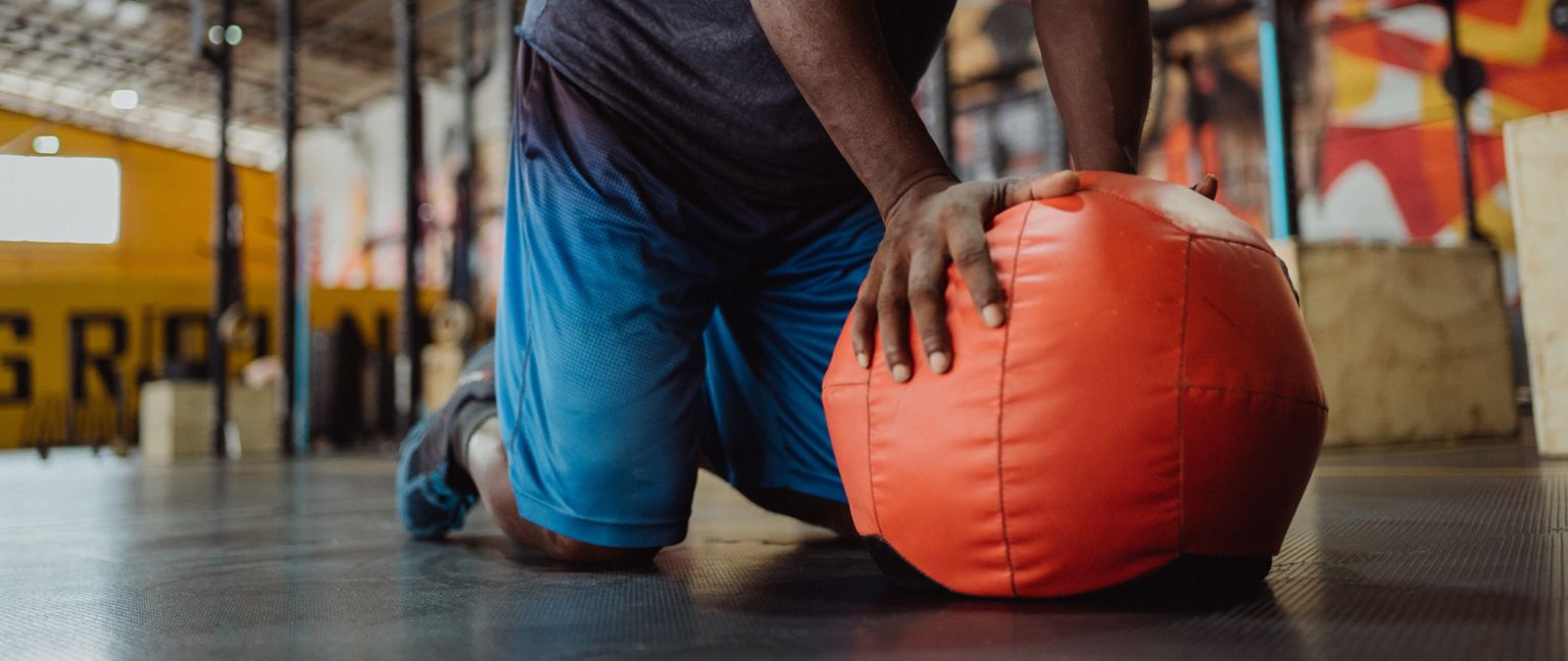 A man in blue shorts and a gray shirt kneels on a gym floor with his hands on an orange wall ball.