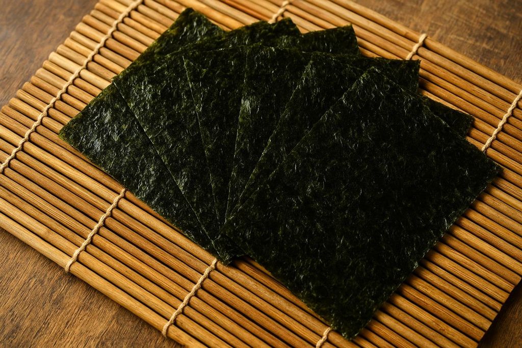 Several sheets of dried, dark green nori seaweed are fanned out on a light brown bamboo rolling mat, with a dark wooden surface visible beneath. The nori sheets have a slightly textured, crinkled appearance.