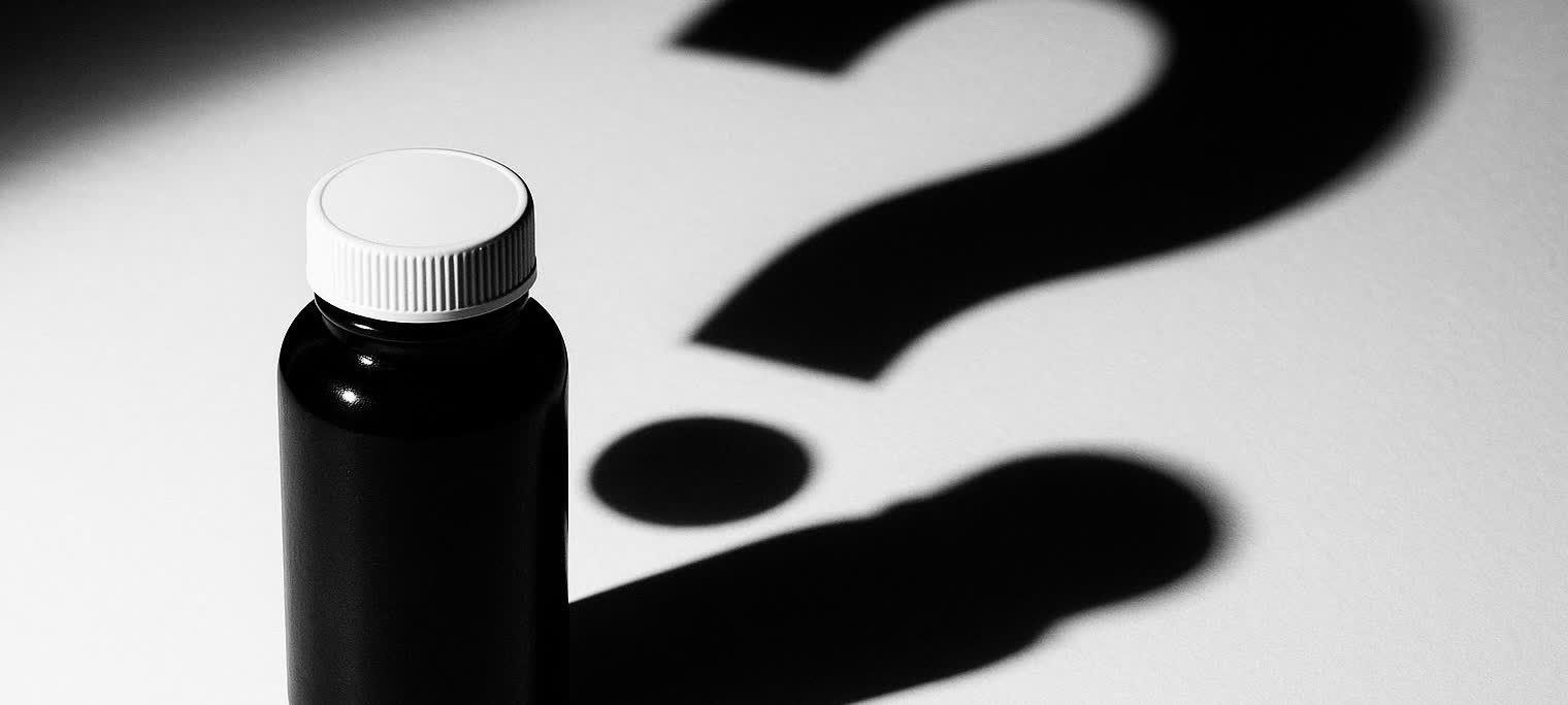 A black medicine bottle with a white cap stands on a white surface, casting a large shadow shaped like a question mark. The image is in black and white.
