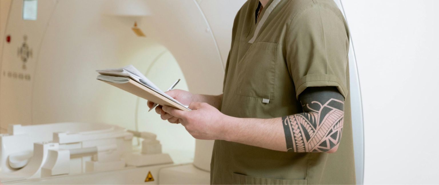 A healthcare professional in olive scrubs stands next to a CT scanner, holding a clipboard and pen in hands with a visible arm tattoo.