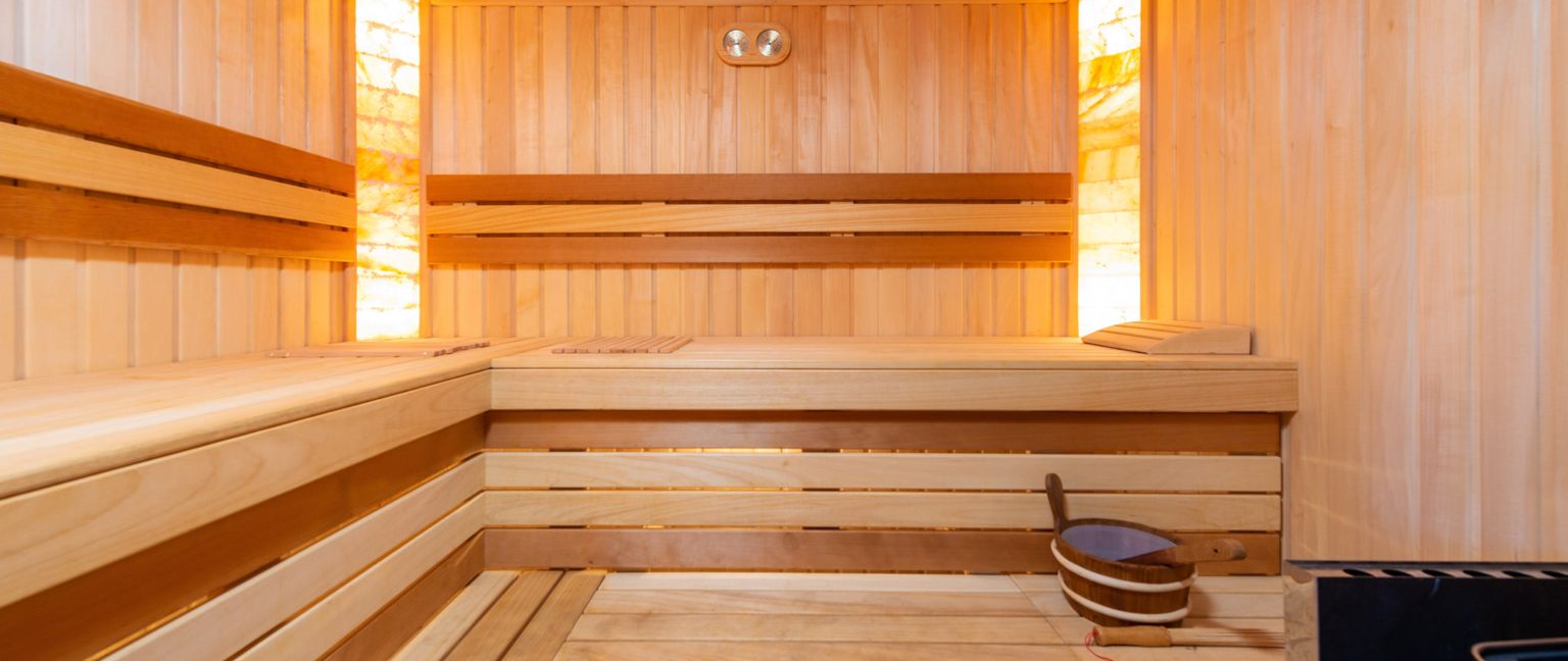 The interior of a modern sauna featuring light wood paneling, tiered benches, illuminated salt panels, and a wooden bucket and ladle on the floor.