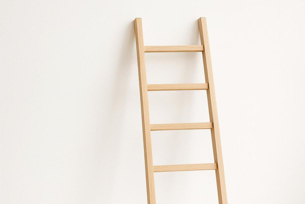 A light-colored, slender wooden ladder leans against a plain white wall. The image features the upper and middle sections of the ladder, highlighting its simple design and smooth texture.