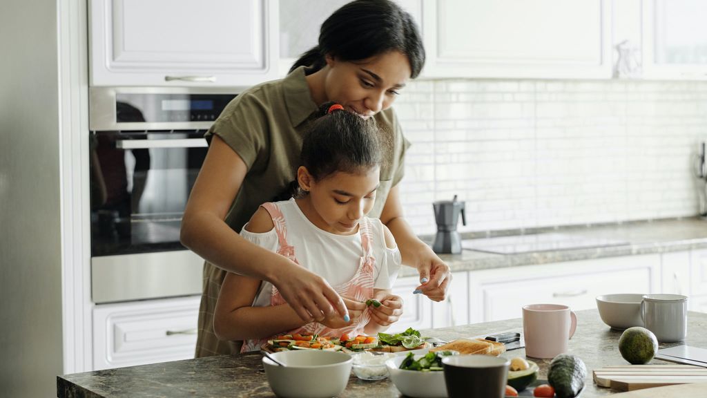 A mother smiles and helps her daughter arrange ingredients on open-faced sandwiches in a sunlit kitchen.