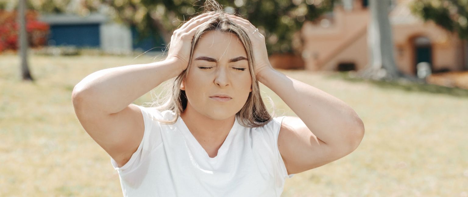 A woman in a white shirt sits outside with her eyes closed, touching her head with both hands as if she has a headache.