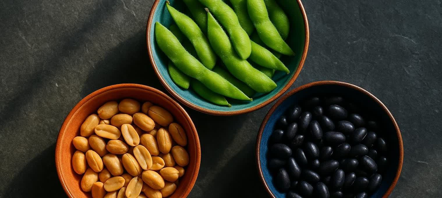 Overhead view of three bowls containing edamame, peanuts, and black beans on a dark background. The edamame are in a blue and orange bowl, the peanuts in an orange bowl, and the black beans in a blue and orange bowl.