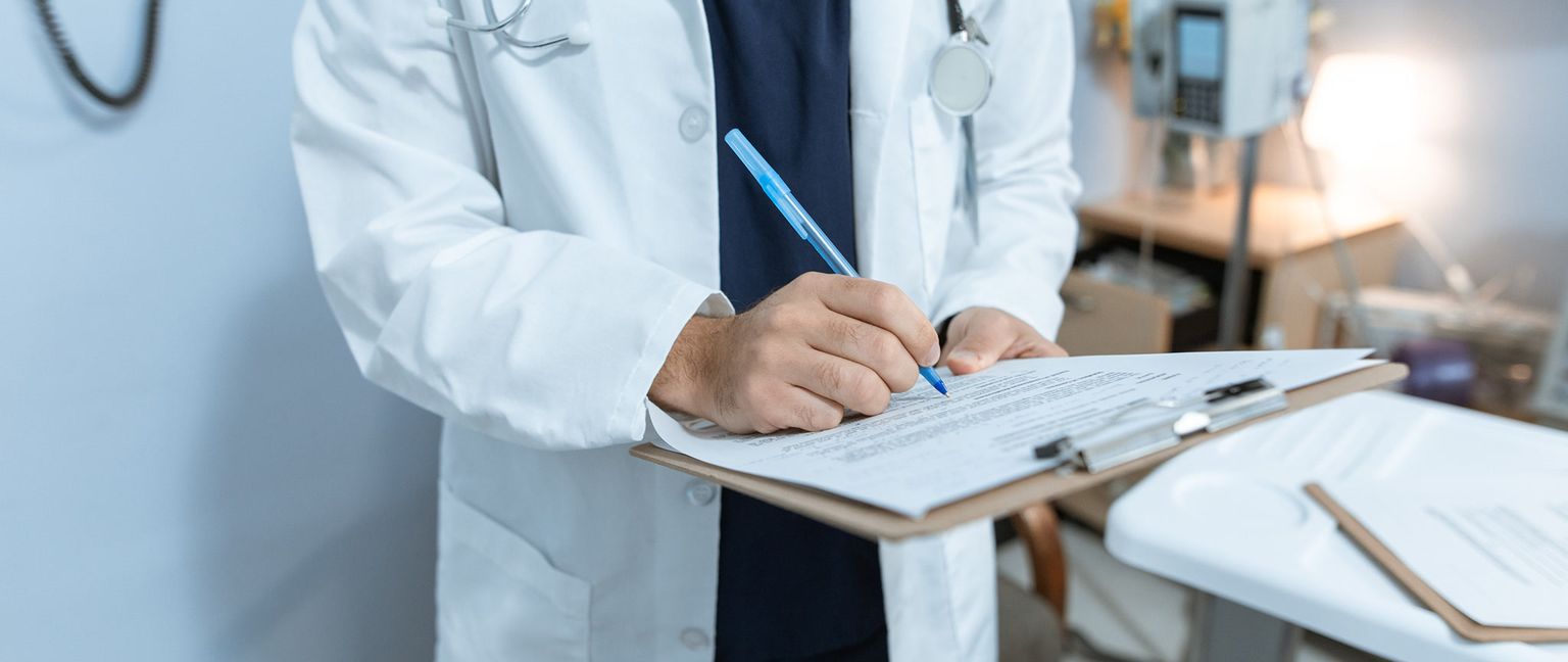 A doctor in a white coat writing with a blue pen on a clipboard with papers.