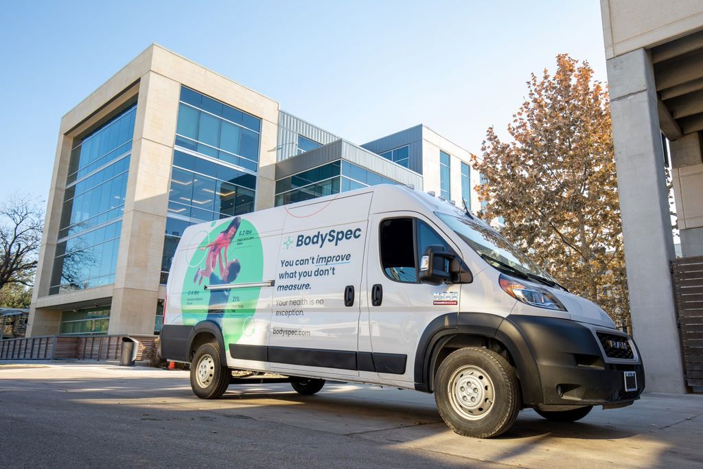 A white BodySpec van with green and teal graphics on its side, including the BodySpec logo and text "You can't improve what you don't measure. Your health is no exception." The van is parked on a paved driveway in front of a modern building with large glass windows and light-colored stone accents. A tree with autumn leaves is visible in the background.