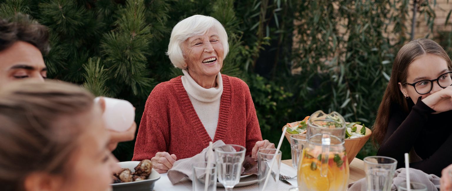 A family gathered around a table outdoors, laughing and enjoying a meal together. The focus is on a senior woman smiling brightly at the center of the frame.