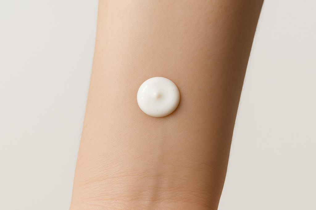 A close-up view of a person's inner forearm with a small, perfectly circular dab of white cream, likely for a patch test, centered in the frame. The skin is smooth and light-toned.