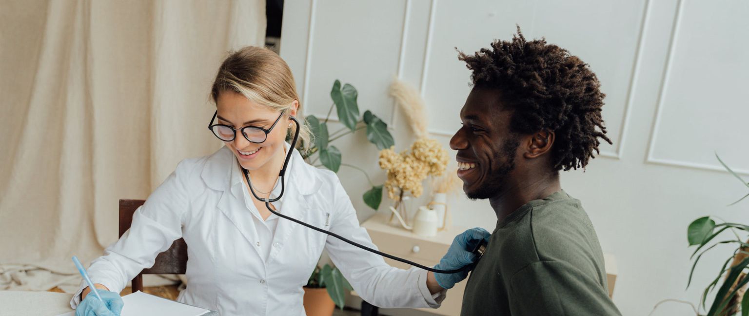 A female doctor in a white coat smiles as she uses a stethoscope to listen to the chest of a male patient, who is also smiling.