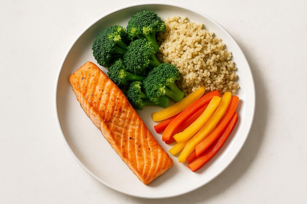 Top-down photo of a healthy meal with salmon, broccoli florets, sliced red and yellow bell peppers, and quinoa on a white plate.