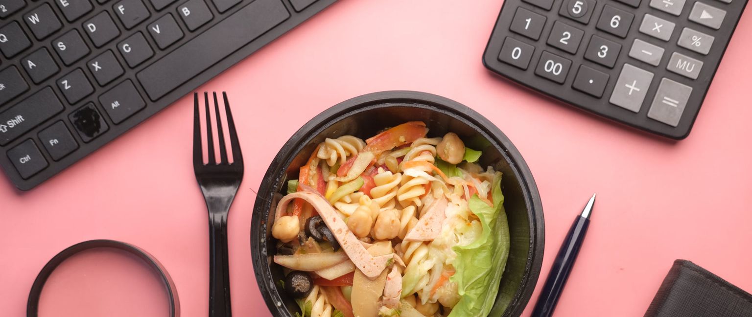 Overhead view of a black calculator, black keyboard, black fork, and black pen and notebook surrounding a bowl of fusilli pasta salad with chickpeas, olives, and lettuce on a pink background.