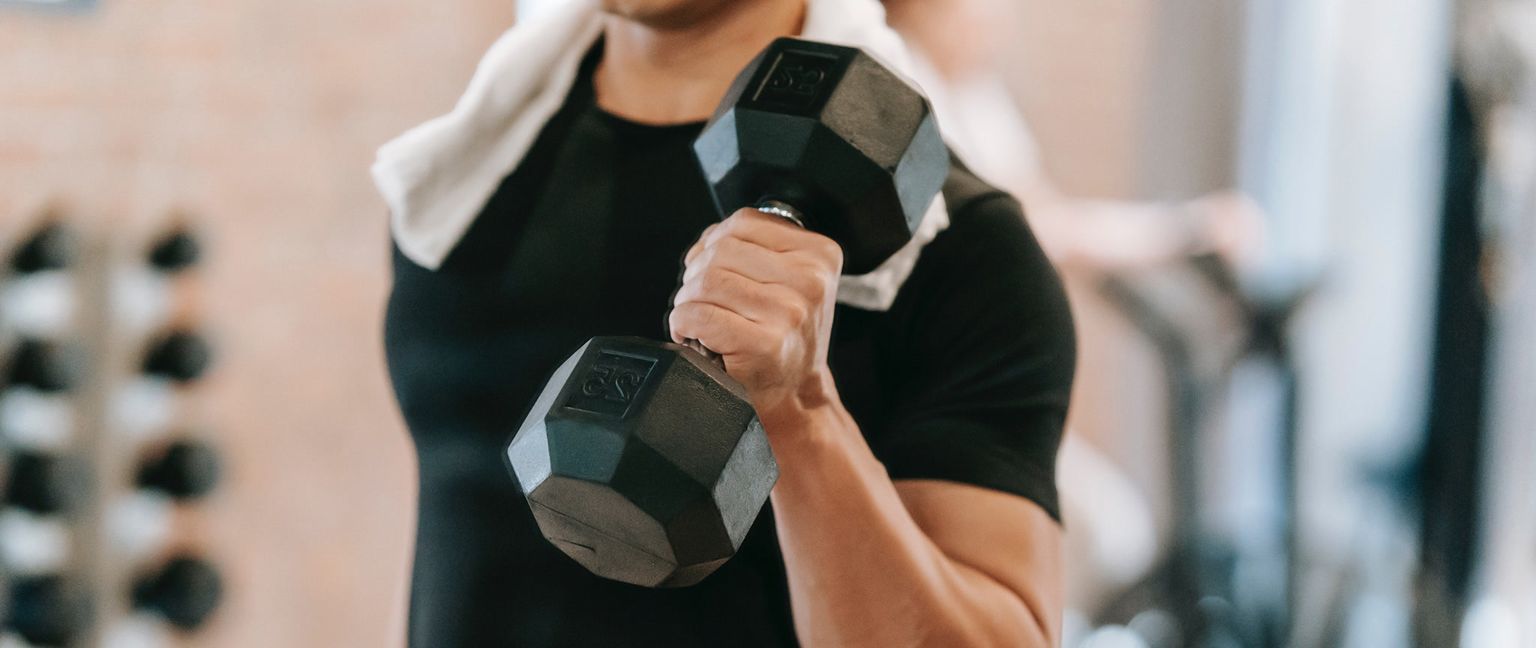 A close-up of a man in a black t-shirt with a white towel around his neck, holding a black dumbbell in his right hand in a gym.