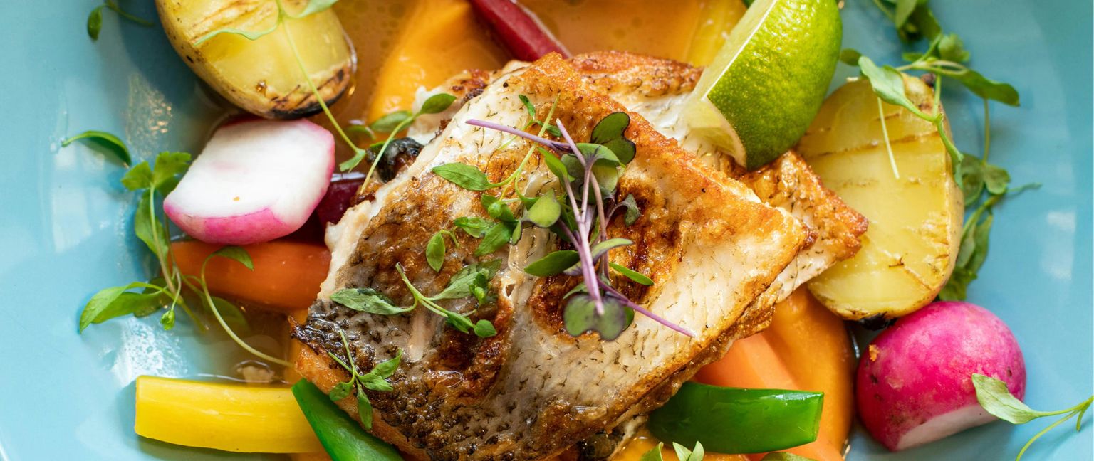 Close-up shot of roasted fish with crispy skin served on a bed of roasted carrots, yellow carrots, and snap peas. The dish is garnished with radishes, grilled potato halves, fresh herbs, and a lime wedge. A light broth is visible in the pale blue bowl.
