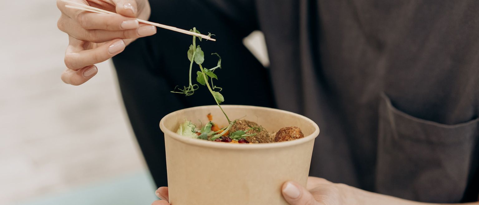 A person picks up a pea shoot with a toothpick from a paper bowl filled with falafel and salad.