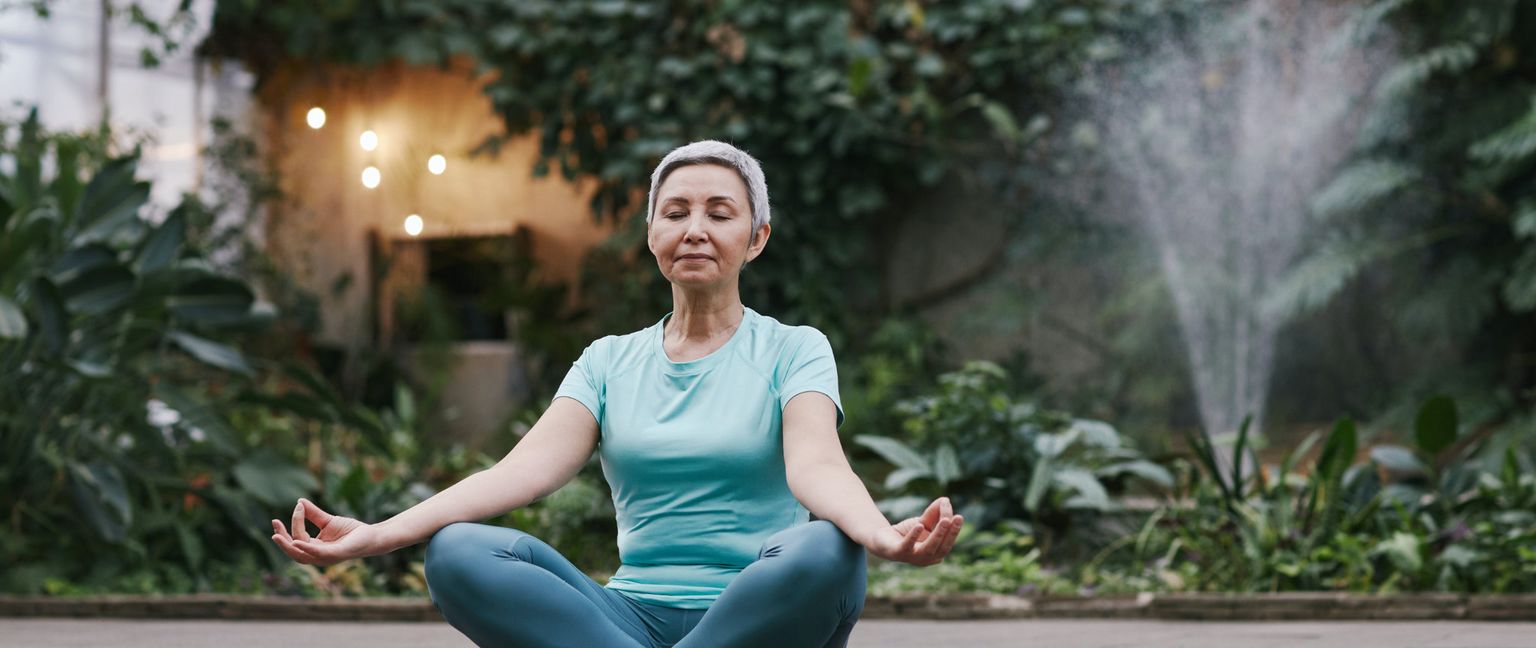 An older woman sits in a cross-legged meditation pose among plants, with a fountain in the background.