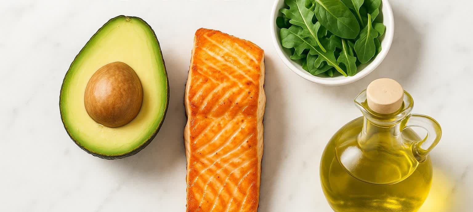 A flat lay of healthy keto diet foods, including a sliced avocado, cooked salmon fillet, a bowl of fresh spinach, and a bottle of olive oil, arranged on a clean white surface with natural light.