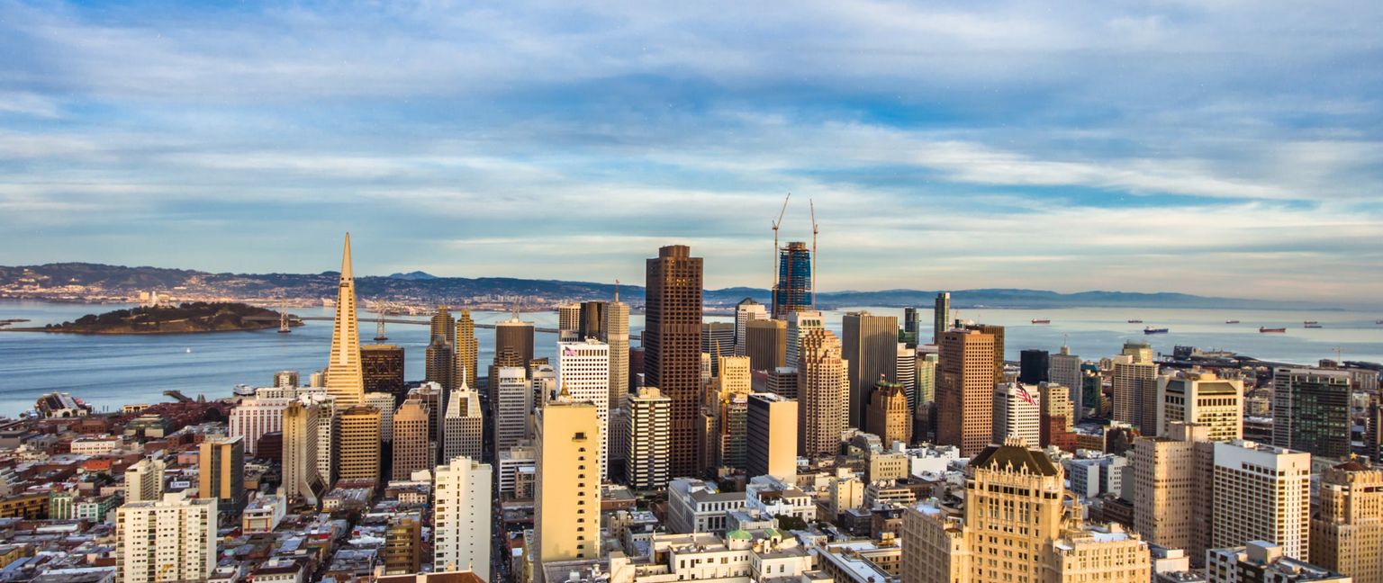 An elevated view of the San Francisco skyline with the bay, Treasure Island, and bridges in the background under a cloudy sky.